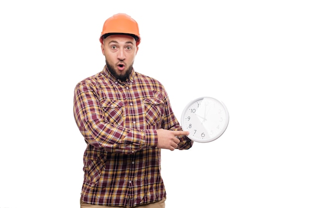 Photo angry and shocked builder worker in protective construction orange helmet holding in hand a big alarm clock isolated on white background. time to work. building construction time.