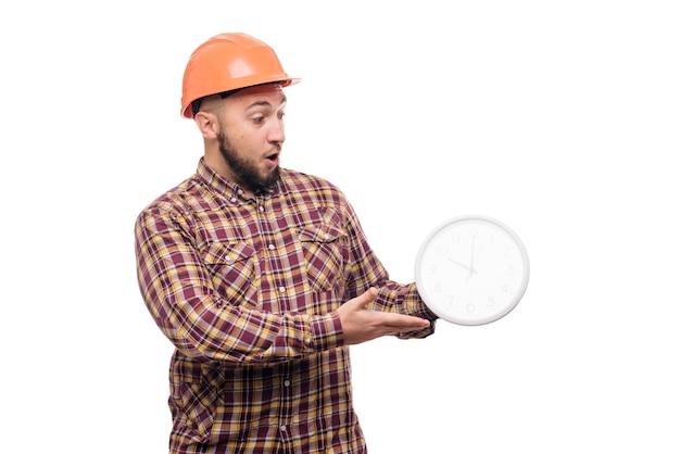 Photo angry and shocked builder worker in protective construction orange helmet holding in hand a big alarm clock isolated on white background. time to work. building construction time.