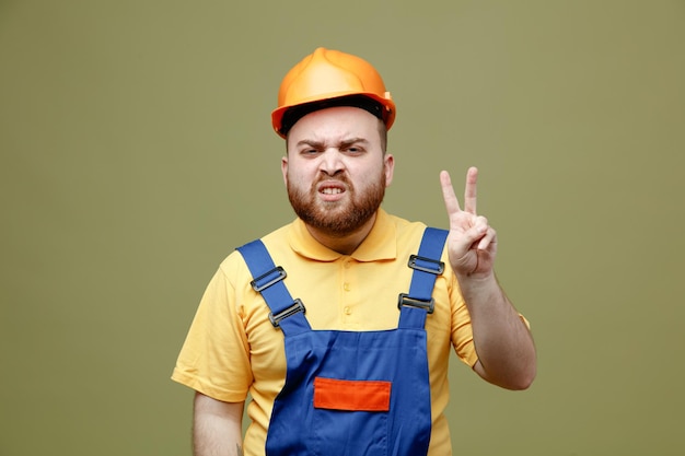 Photo angry showing peace gesture young builder man in uniform isolated on green background