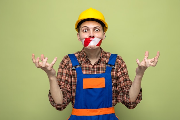 Angry spreading hands young male builder wearing uniform sealed mouth with duct tape 