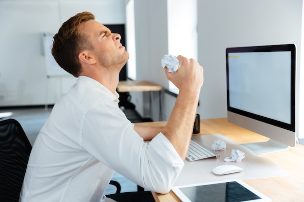 Angry stressed young businessman sitting and crumpling paper in office