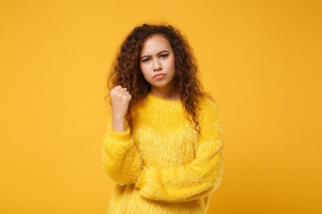 Angry strict young african american girl in fur sweater posing isolated on yellow orange background, studio portrait. People sincere emotions lifestyle concept. Mock up copy space. Clenching fists.