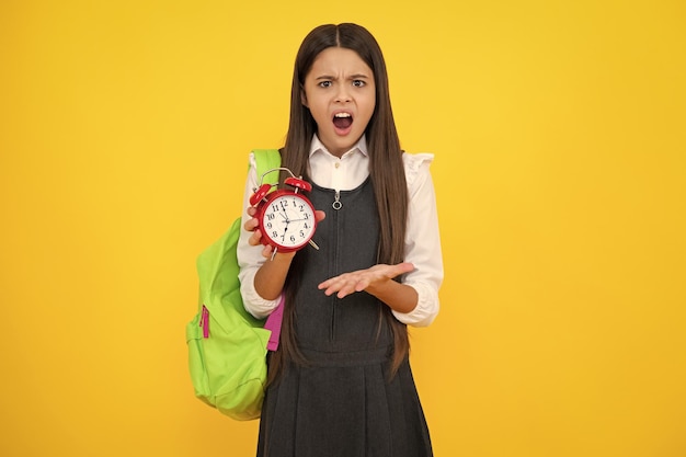 Photo angry teenager girl upset and unhappy negative emotion back to school teenager school girl with backpack hold clock alarm time to learn school children on isolated yellow background