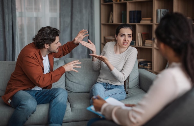 Angry unhappy millennial european husband and wife arguing at consultation with black doctor in