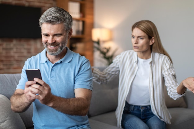 Angry upset adult european wife yelling at smiling husband with smartphone in living room interior