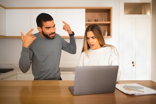 Angry upset millennium arab guy and european female look at laptop quarreling from finance and debt