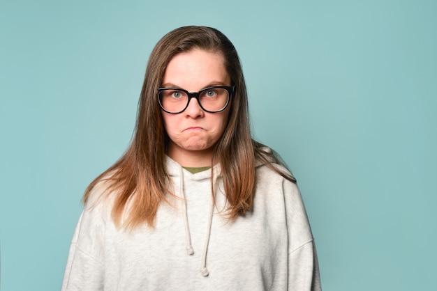 Angry and upset student girl wearing glasses and a hoodie on a teal wall