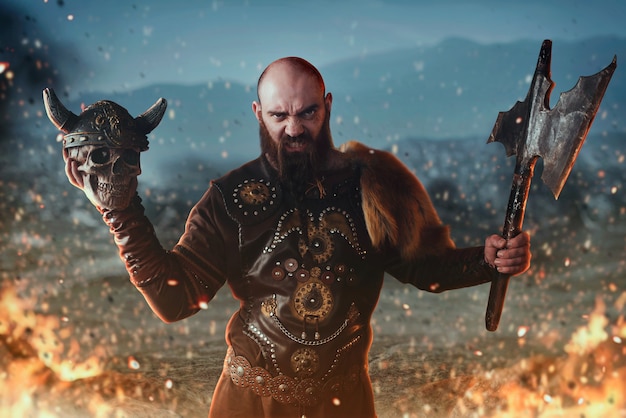 伝統的な北欧の服を着た怒っているバイキングは、斧と人間の頭蓋骨を保持し、火の中で戦います。スカンジナビアの古代の戦士