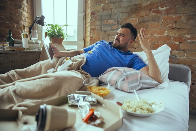 Angry watching tablet Lazy caucasian man living in his bed surrounded with messy