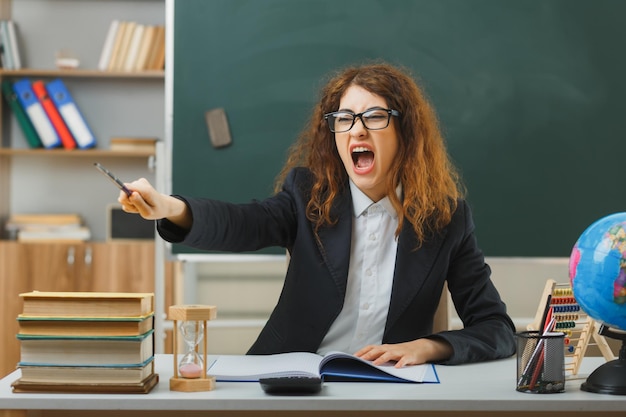 angry with closed eyes young female teacher wearing glasses points at camera with pointer sitting at desk with school tools in classroom