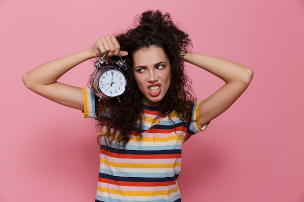 angry woman 20s with curly hair holding alarm clock isolated on pink