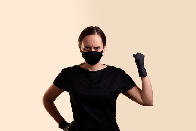 Сердитая женщина в черной маске для лица и латексных перчатках показывает кулак над бежевой стеной