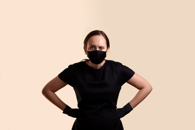 Angry woman in black dress face mask and latex gloves standing with arms akimbo and  over beige wall