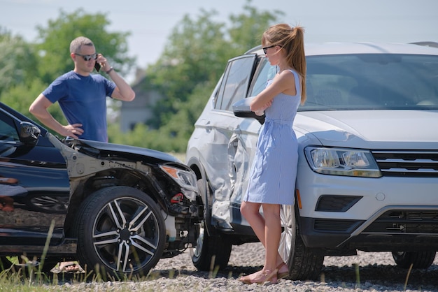 Angry woman and man drivers of heavily damaged vehicles calling\
insurance service for help in car crash accident on street side\
road safety concept