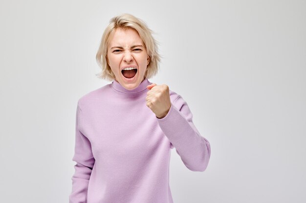 Angry woman in a purple sweater clenching her fists and shouting isolated on a white background