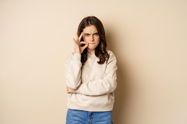 Angry woman shows mouth zipper, seal lips gesture, standing\
over beige background, taboo sign