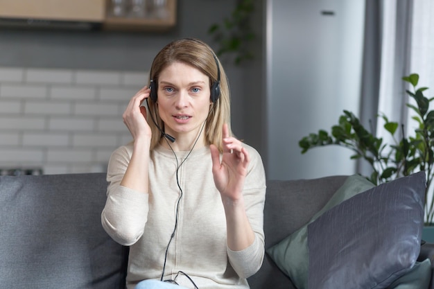 Photo angry woman talking on video call looks at camera and explains uses headset works remotely from home sitting on sofa