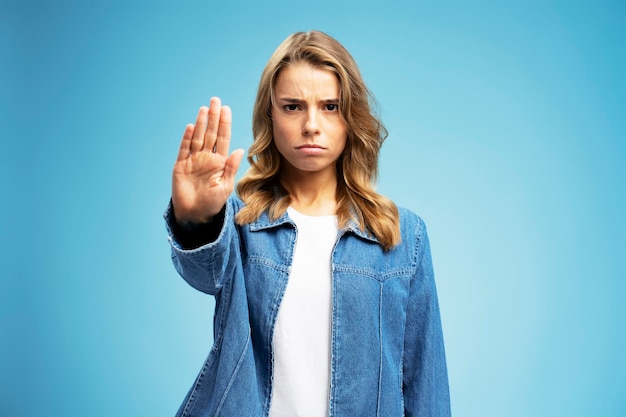 angry woman wearing casual denim clothes showing stop sign looking at camera isolated on blue