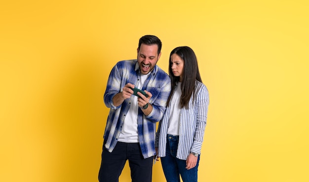 Angry young adult girlfriend looking at cheerful boyfriend screaming ecstatically and playing video game on smart phone isolated over yellow background