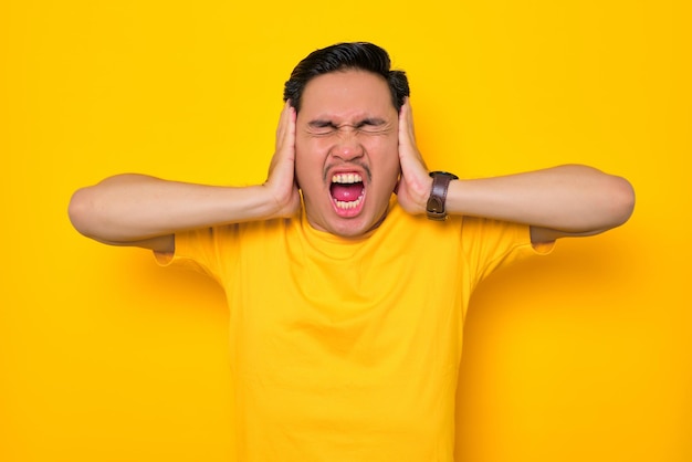 Angry young Asian man in casual tshirt screaming with closed eyes and open mouth covering ears with hands isolated on yellow background People lifestyle concept