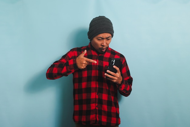 Angry young Asian man is yelling and pointing at his phone isolated on a blue background