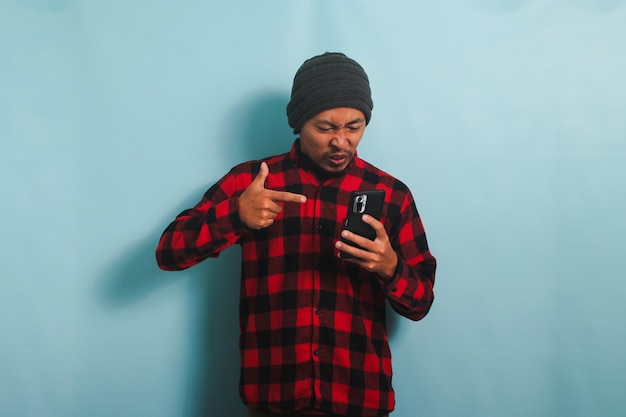 Photo angry young asian man is yelling and pointing at his phone isolated on a blue background