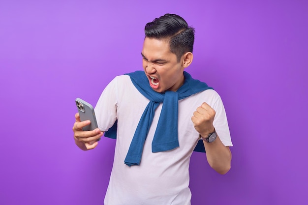 Angry young Asian man using mobile phone reacting to fake news isolated over purple background