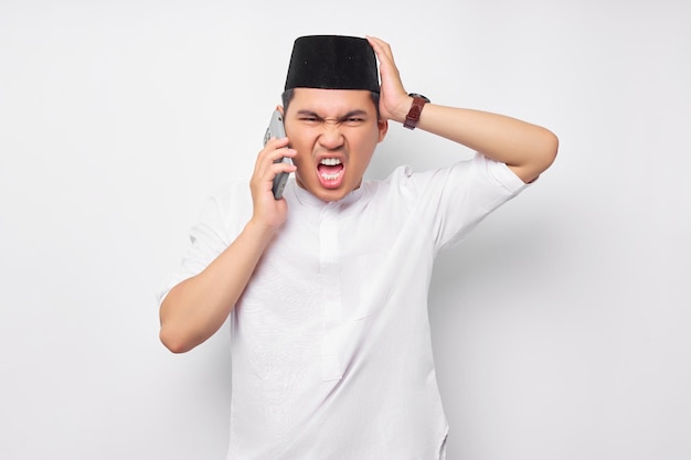 Angry young Asian Muslim man talking on mobile phone keeps hands on head isolated on white background People religious Islam lifestyle concept celebration Ramadan and ied Mubarak