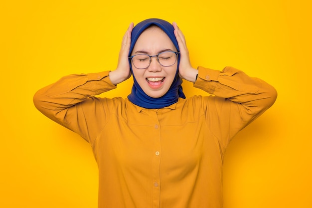 Angry young Asian Muslim woman dressed in orange covering ears with hands and screaming loudly isolated on yellow background