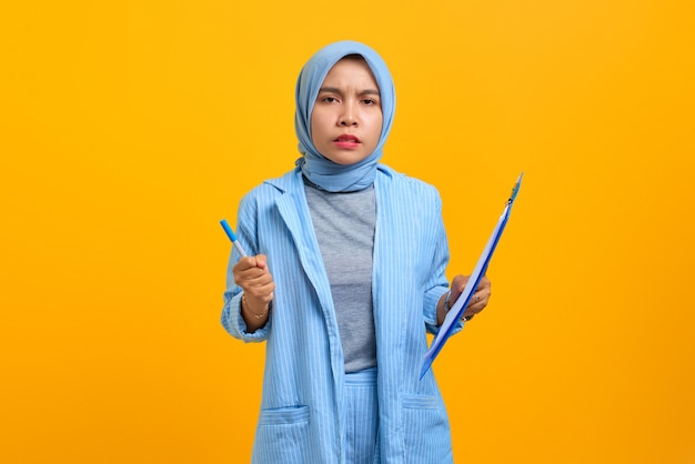 Angry young asian woman holding pen and document folder over yellow background