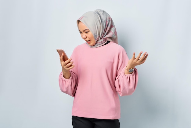 Angry young asian woman in pink shirt using mobile phone while\
loudly shouting isolated over white background