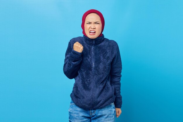 Angry young Asian woman screaming out with raised hand isolated on blue background