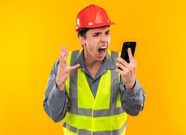 Photo angry young builder man in uniform holding and looking at phone