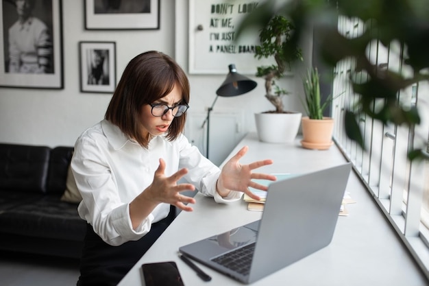 Angry young businesswoman working online on laptop getting video call or reading emails sitting at