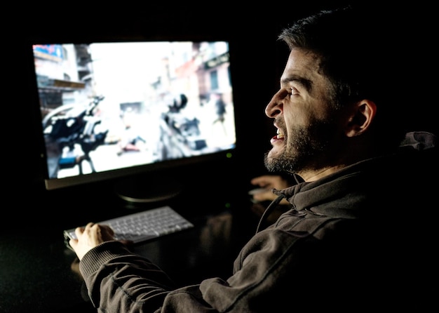 Разгневанный молодой игрок играет в видеоигры на компьютере в темной комнате