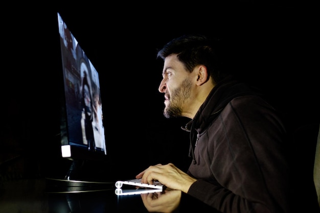 Angry young gamer playing videogame on computer in dark room