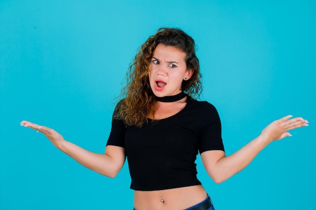 Angry young girl is looking down by opening wide her hands on blue background