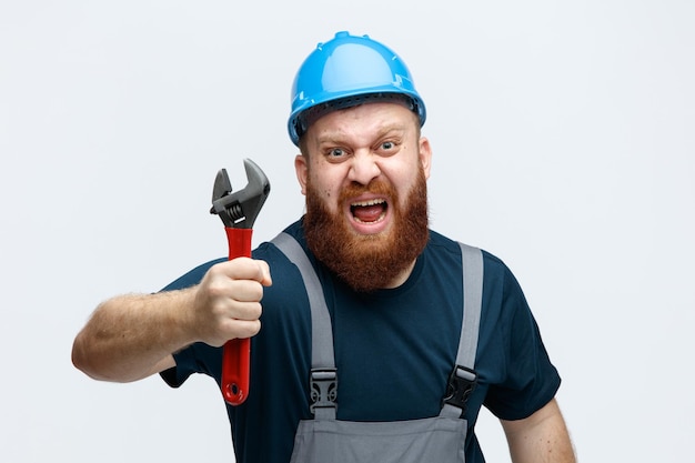 Злой молодой мужчина-строитель в защитном шлеме и униформе смотрит в камеру, держа гаечный ключ, крича на белом фоне