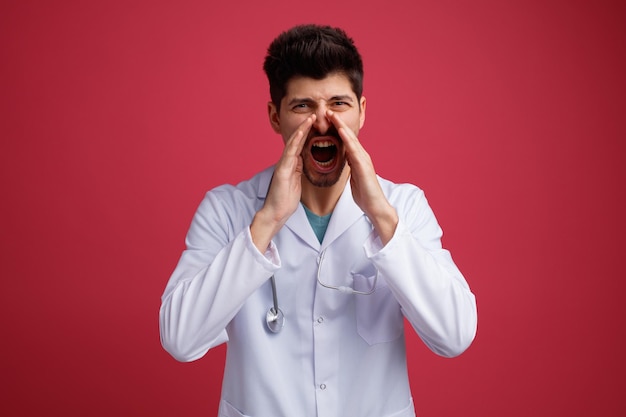 Angry young male doctor wearing medical uniform and stethoscope around his neck looking at camera keeping hands near mouth shouting out loud something isolated on red background