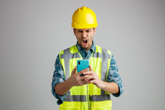 Angry young male engineer wearing safety helmet and uniform holding and looking at mobile phone shouting isolated on white background