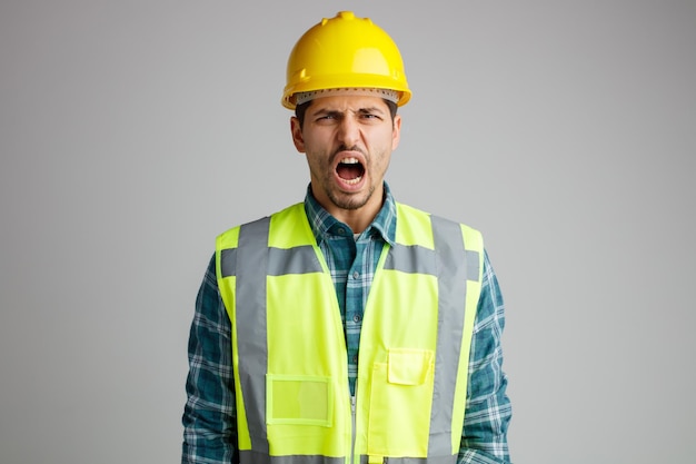 Angry young male engineer wearing safety helmet and uniform looking at camera screaming isolated on white background