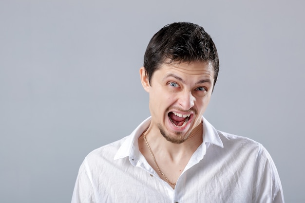 Angry young man brunette in the white shirt screaming on a grey background.