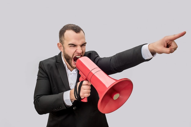 Angry young man hold red megaphone in hand nad screma into it. He points forward. Guy is mad. 