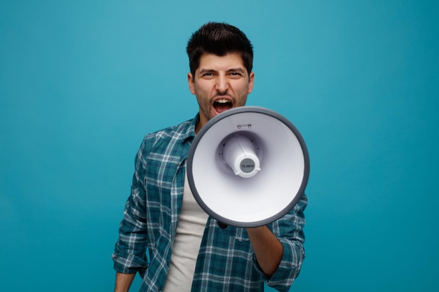 Angry young man looking at camera talking into speaker isolated on blue background