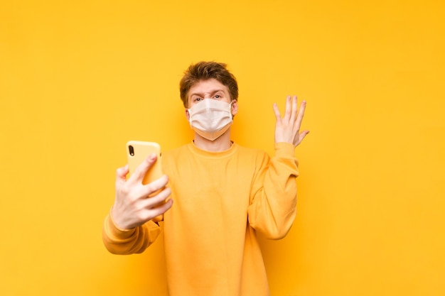 黄色の背景に彼の手でスマートフォンで隔離の医療マスクで怒っている若い男