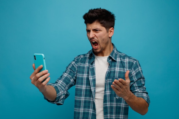 Angry young man stretching mobile phone out looking at it keeping hand in air shouting isolated on blue background