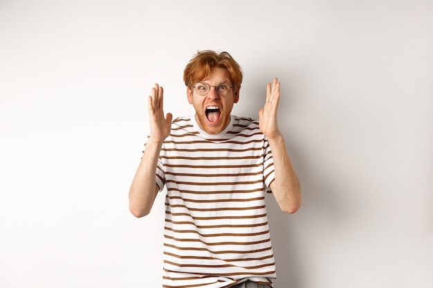 Angry young man with red hair shouting at camera, screaming and looking outraged, shaking hands, standing over white background.