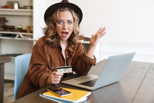 angry young woman in hat expressing outrage and holding credit card while working with laptop