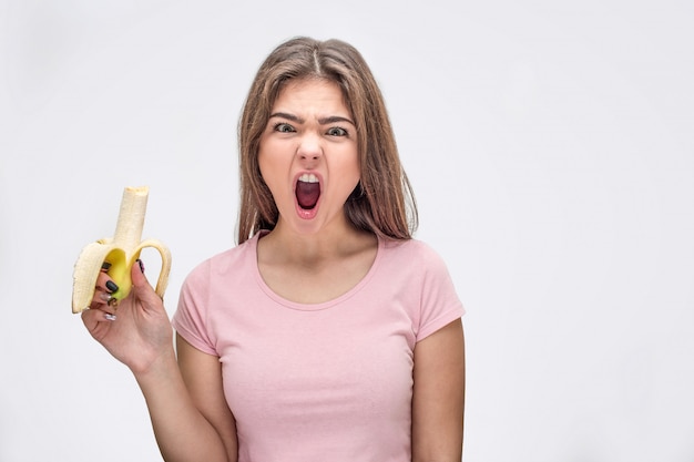 Angry young woman looks at the camera and screams while holding a banana