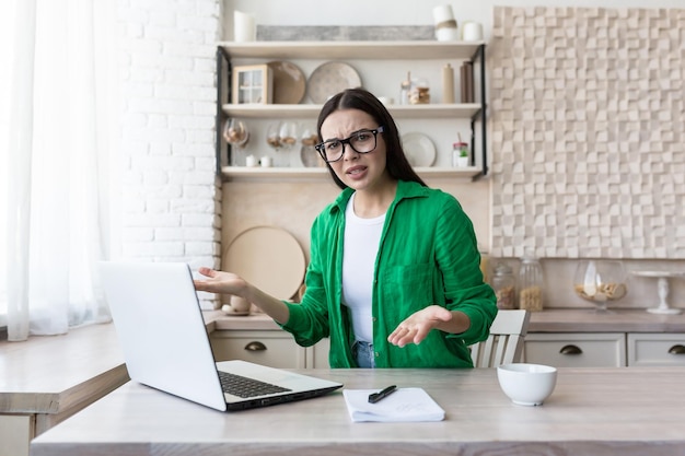 Angry young woman sitting at home at kitchen table in front of laptop with notebook and documents he